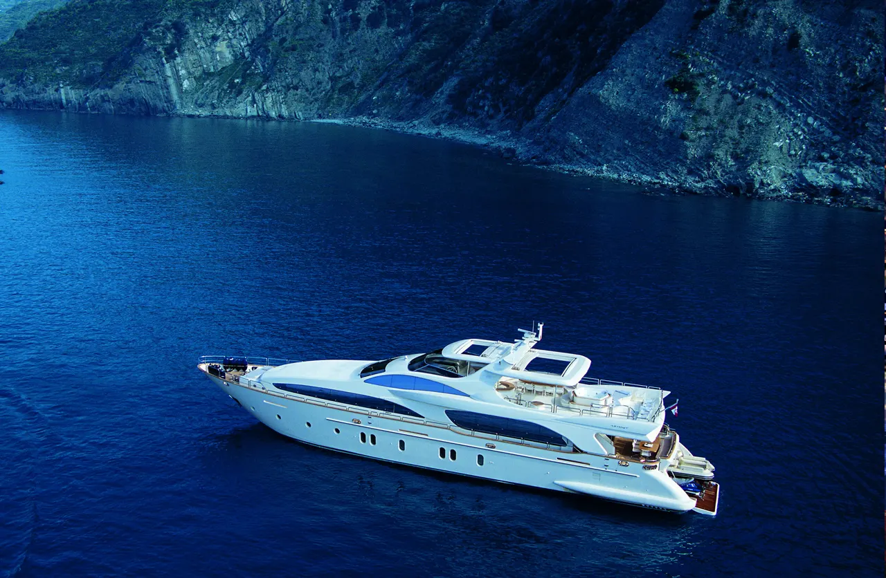 A motor yacht underway in deep blue water near a rocky coastline, seen from above